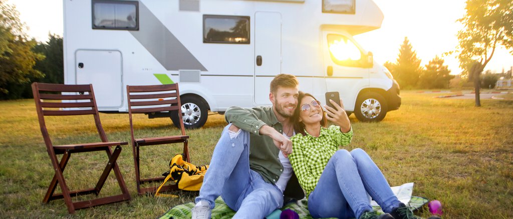 Auf dem Bild sitzen ein Mann und eine Frau auf einer Picknickdecke vor ihrem Wohnmobil und campen sorgenfrei dank der Wohnmobil-Versicherung und Wohnwagen-Versicherung.
