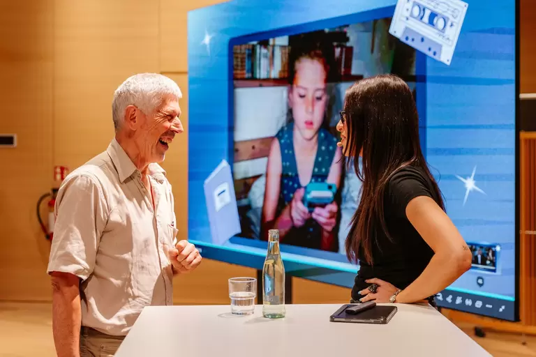 Salwa Houmsi im Gespräch mit einem älteren Herren, vor einem Bildschirm auf dem ein Kinderfoto zu sehen ist