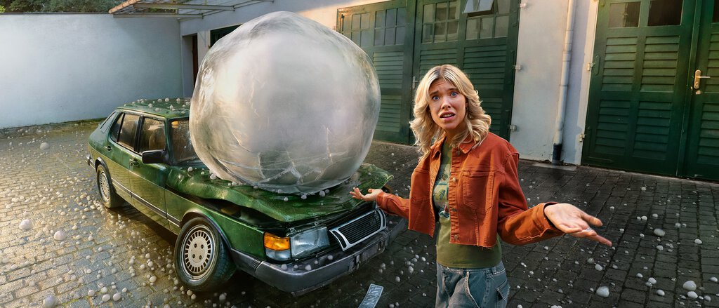 Eine Frau steht vor einem Auto auf das ein überdimensional großes Hagelkorn gefallen ist