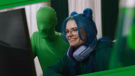 Ein Mädchen mit blauen Haaren ist am zocken, hinter ihr steht Larry, das LVM-Gaming-Maskottchen in einen grünen Ganzkörperanzug.