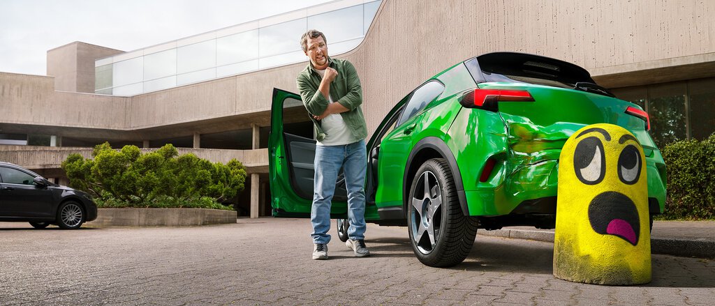 Humorvolles Bild: Ein junger Mann steht fassungslos neben einem grünen Kleinwagen und betrachtet das zerbeulte Heckteil. Er ist beim Rückwärtsfahren mit Schwung gegen einen dicken Betonpfosten gefahren. Der Pfosten ist knallgelb angestrichen und zeigt ein comicartiges empörtes Gesicht.