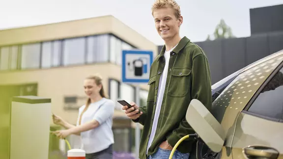 Ein junger Mann schaut auf sein Smartphone während er sein E-Auto lädt