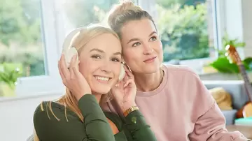 Zwei junge Frauen sitzen auf der Couch im stylischen Wohnzimmer und hören mit Kopfhörer Musik.