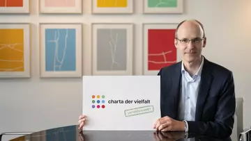 LVM-Vorstandsvorsitzender Dr. Mathias Kleuker hält ein Plakat mit der Aufschrift Charta der Vielfalt in Händen, sitzend vor einer Wand mit Kunstwerken