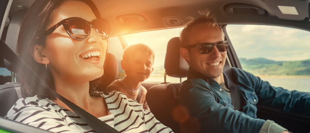 Eine Frau und ein Mann sitzen mit ihrem Kind im Auto und fahren lachend und unbesorgt dank der Kfz-Versicherung für Zusatzfahrer, Mietwagen, Probefahrten und Carsharing.