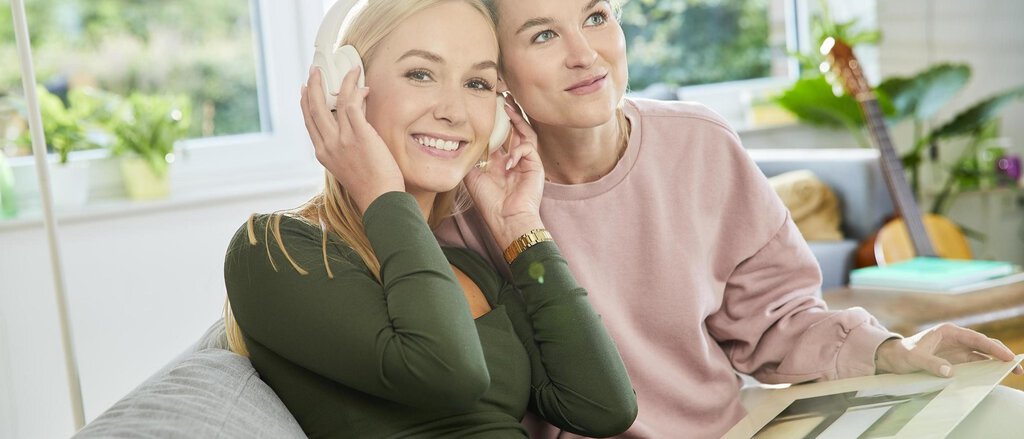 Zwei junge Frauen sitzen auf der Couch im stylischen Wohnzimmer und hören mit Kopfhörer Musik.