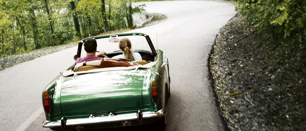 Oldtimer-Versicherung Ein Mann und eine Frau fahren in ihrem grünen Oldtimer dank der Oldtimerversicherung sorgenfrei durch einen Wald.
