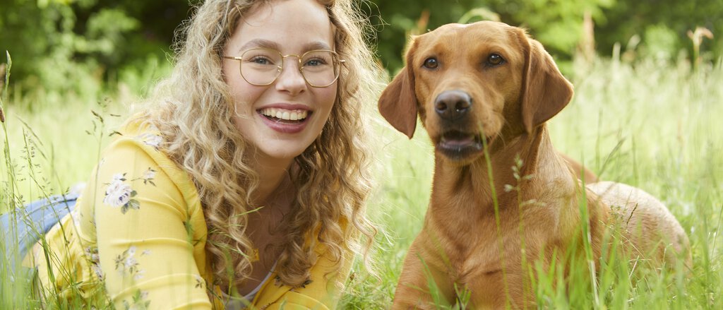 Hundekrankenversicherung Junge Frau liegt mit Hund auf Wiese und lächelt